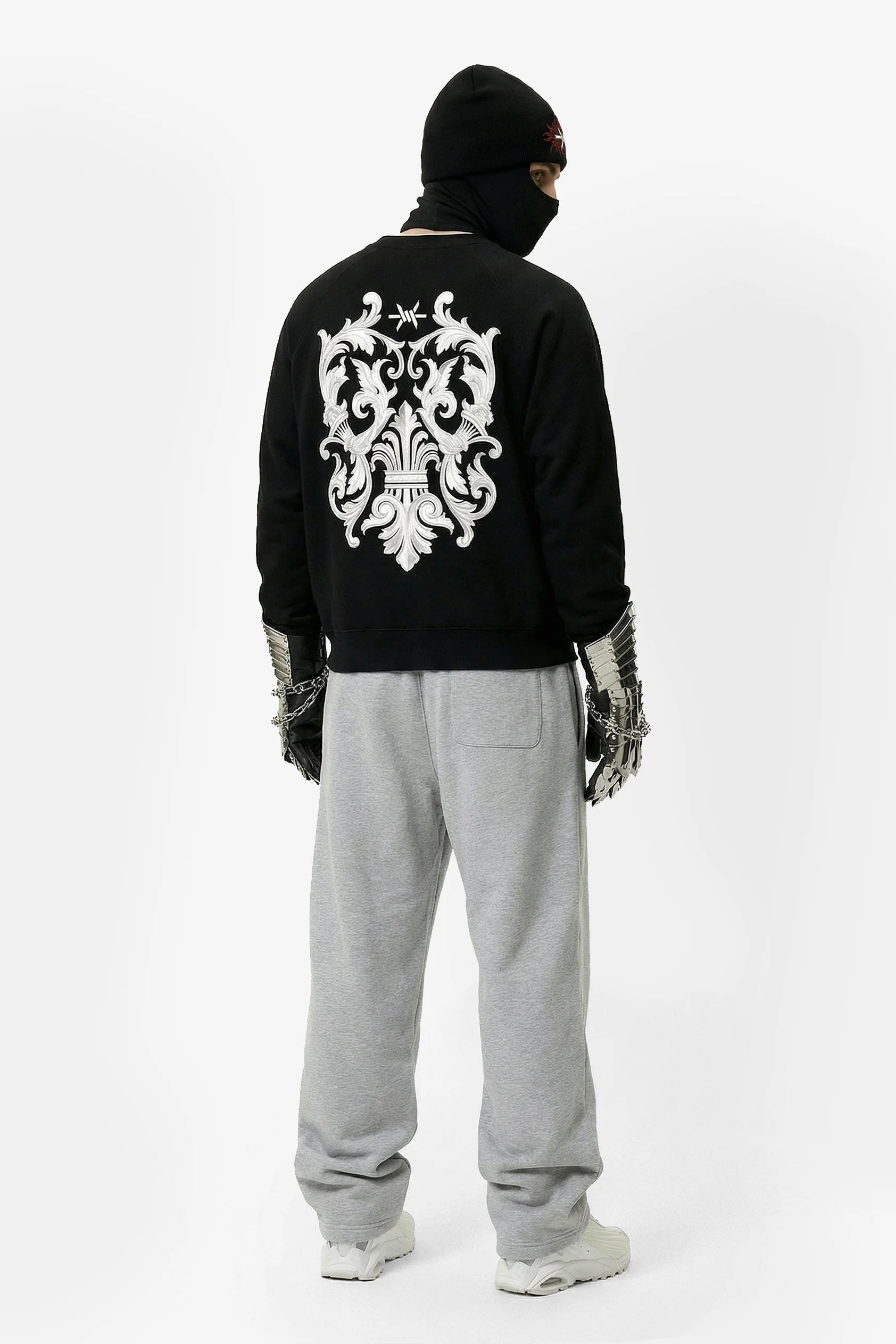 Person wearing a black hoodie with a white emblem on the back, gray pants, and white shoes on a white background.