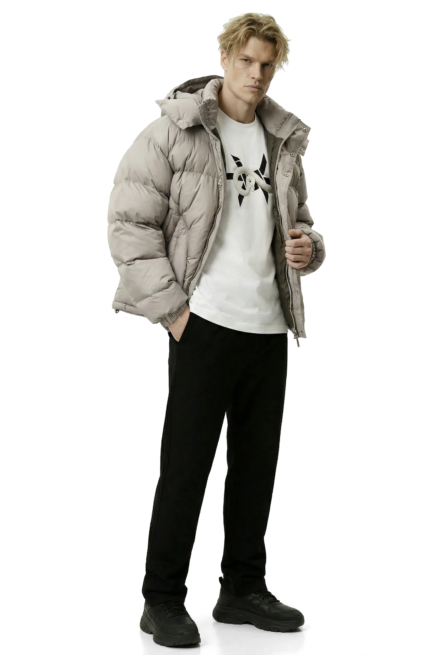Man wearing a light gray khaki puffer jacket, white t-shirt with black design, and black pants on a white background