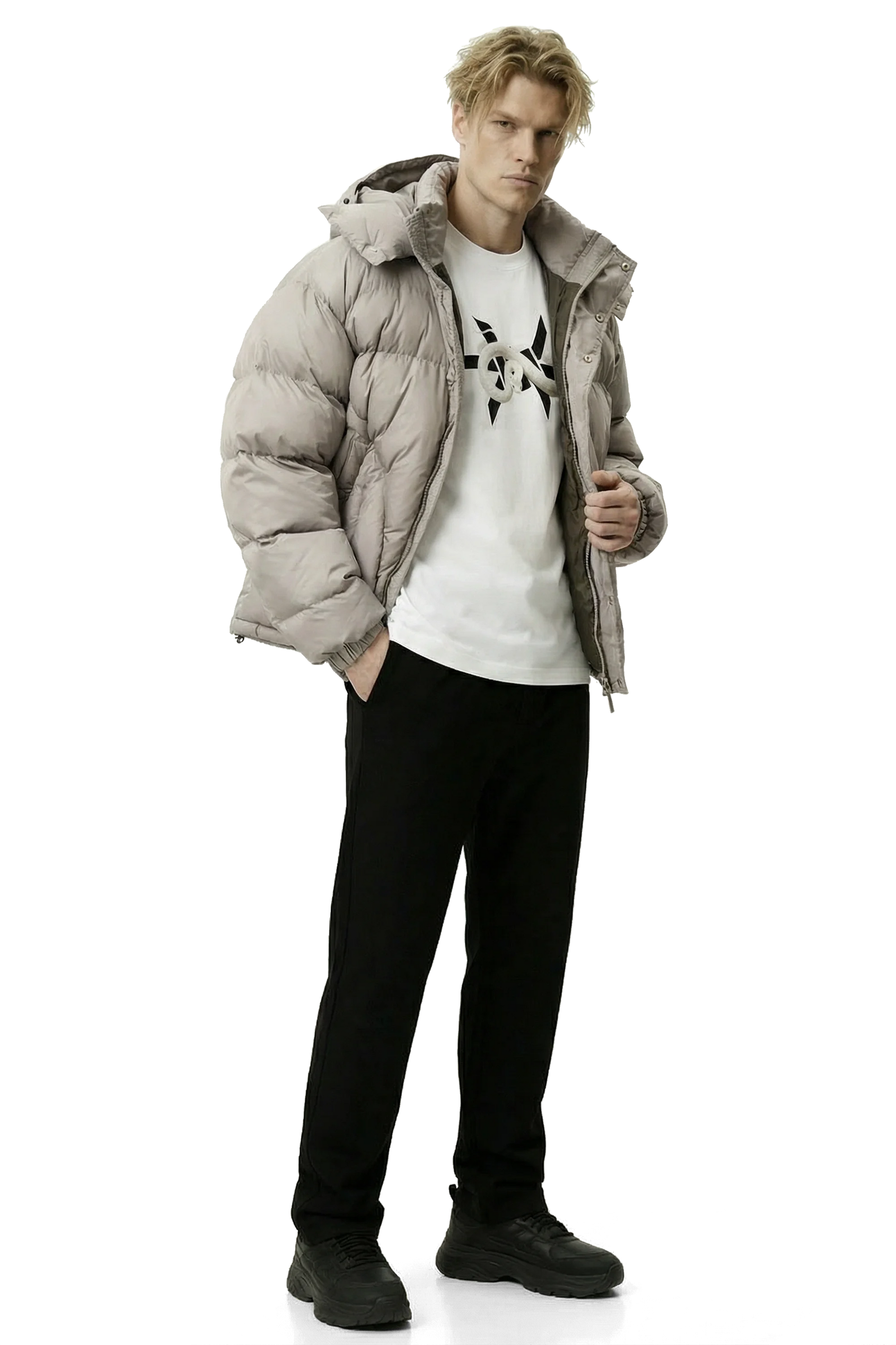 Man wearing a light gray khaki puffer jacket, white t-shirt with black design, and black pants on a white background