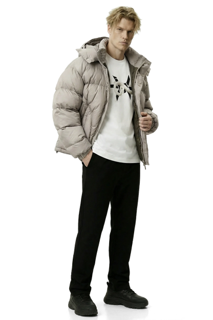 Man wearing a light gray khaki puffer jacket, white t-shirt with black design, and black pants on a white background