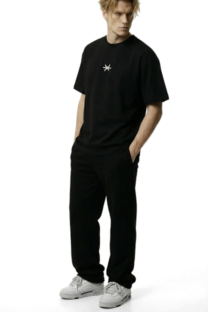Ominous Ascendance Heavyweight Tee - Man wearing an oversized heavyweight black t-shirt with a small white 'Ominous' logo in the upper centre chest area and black pants on a white background