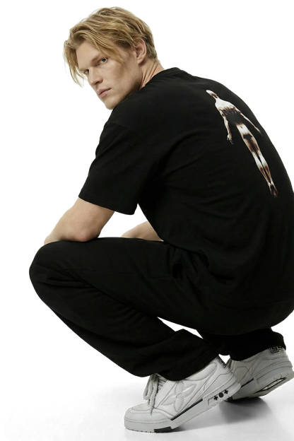 Ominous Ascendance Heavyweight Tee - Strawberry blonde hair white man wearing a black t-shirt with a floating ascending man design on the back with black sweatpants with light gray sneakers on a white background