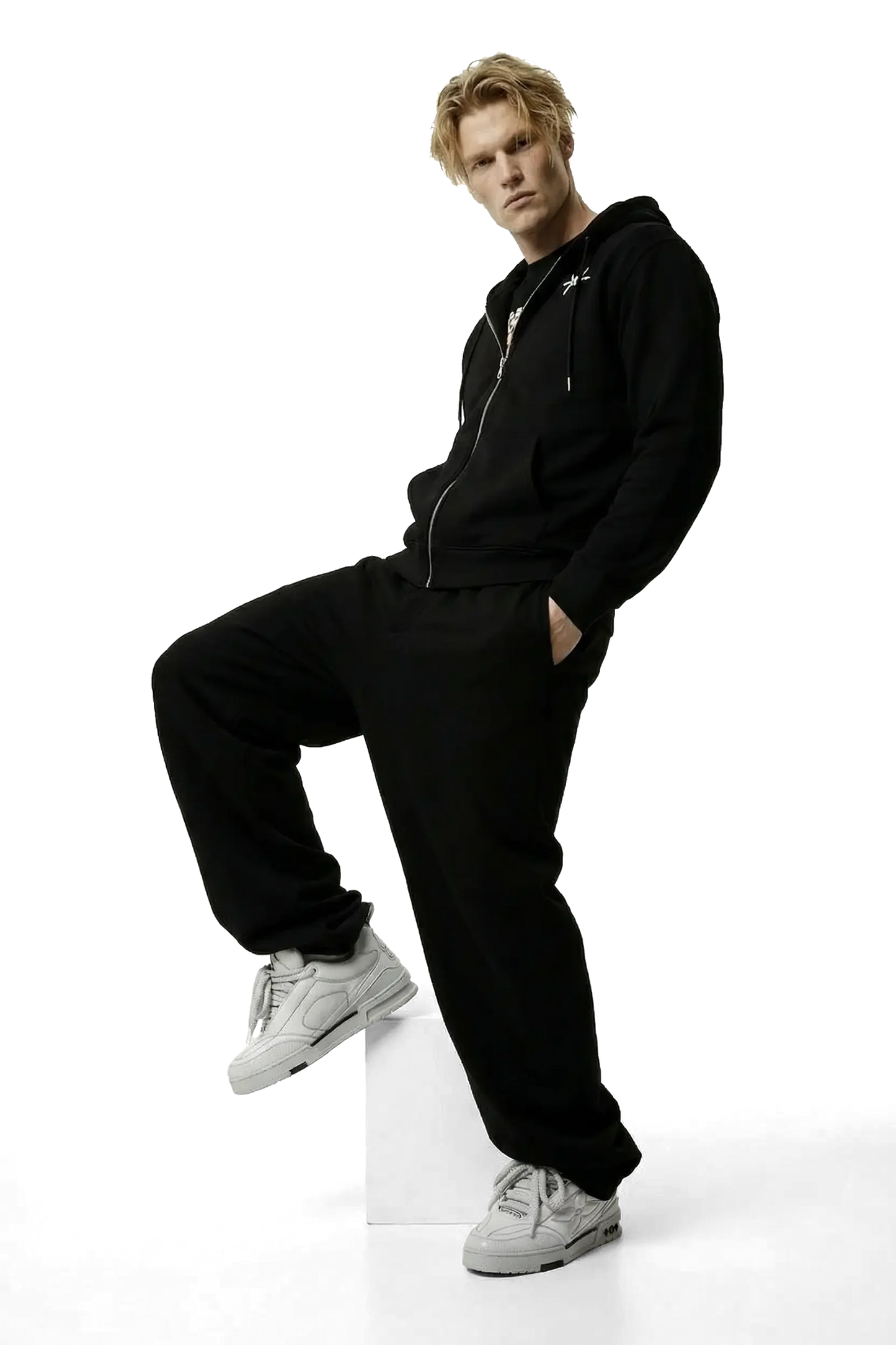 Person wearing a black hoodie and pants with white sneakers on a white background posing on a box