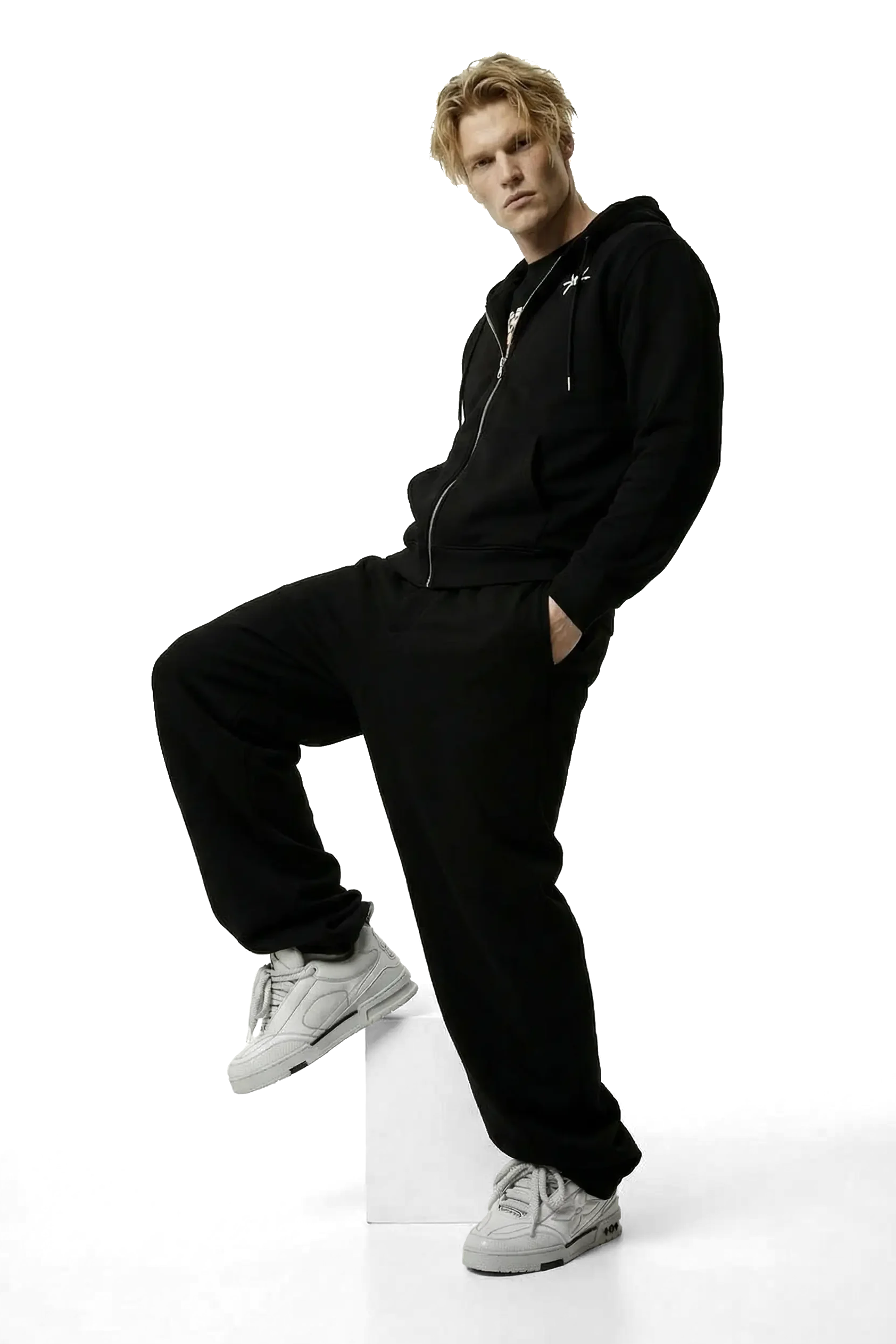 Person wearing a black hoodie and pants with white sneakers on a white background posing on a box