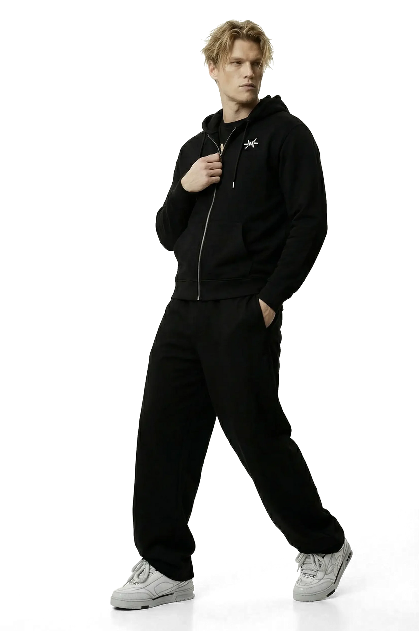 Man wearing a black zip up hoodie with a small logo and black trousers on a white background