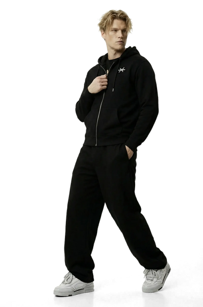 Man wearing a black zip up hoodie with a small logo and black trousers on a white background