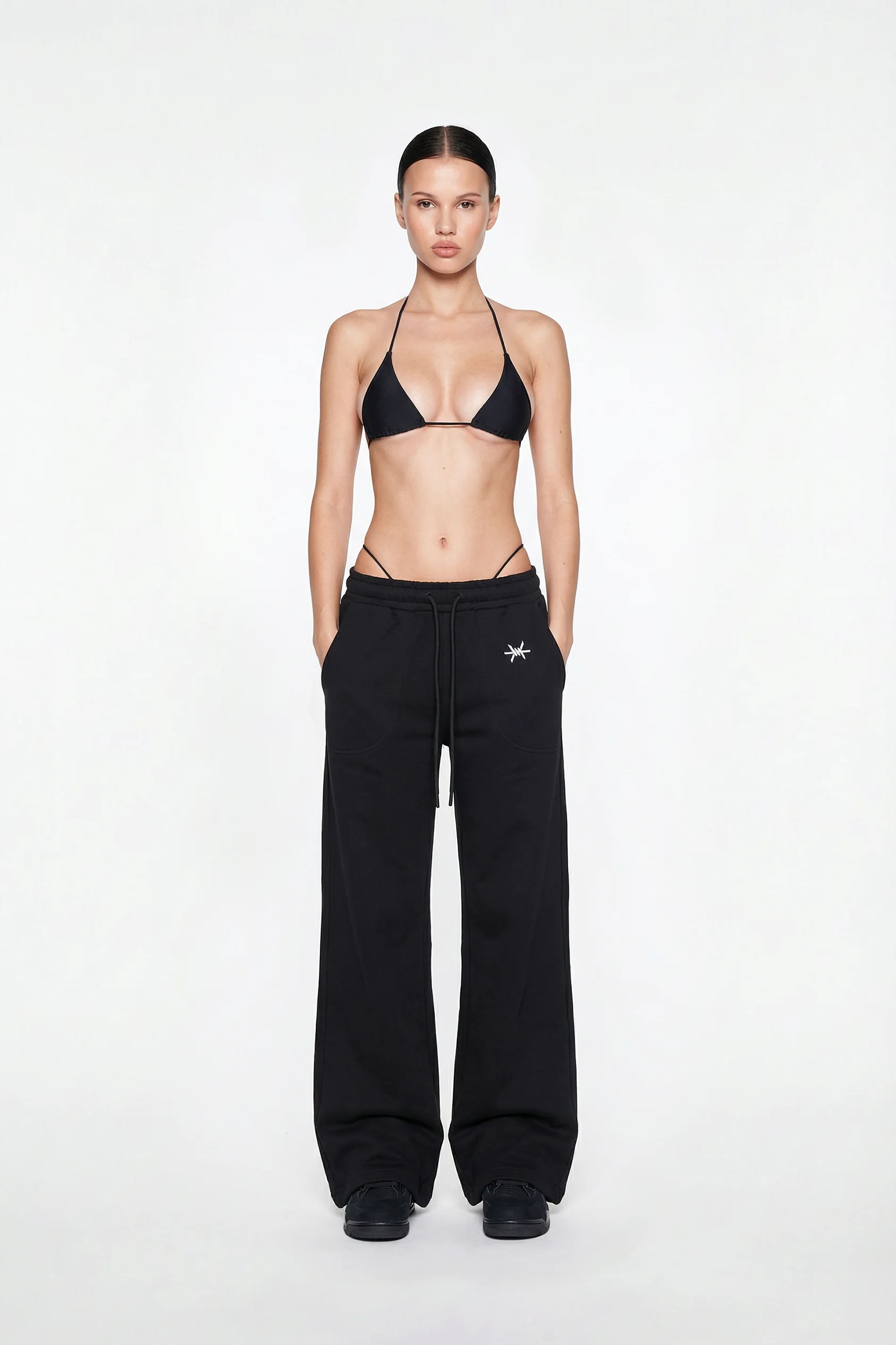 Model wearing black high-waisted pants with a small logo and a black bikini top on a white background.