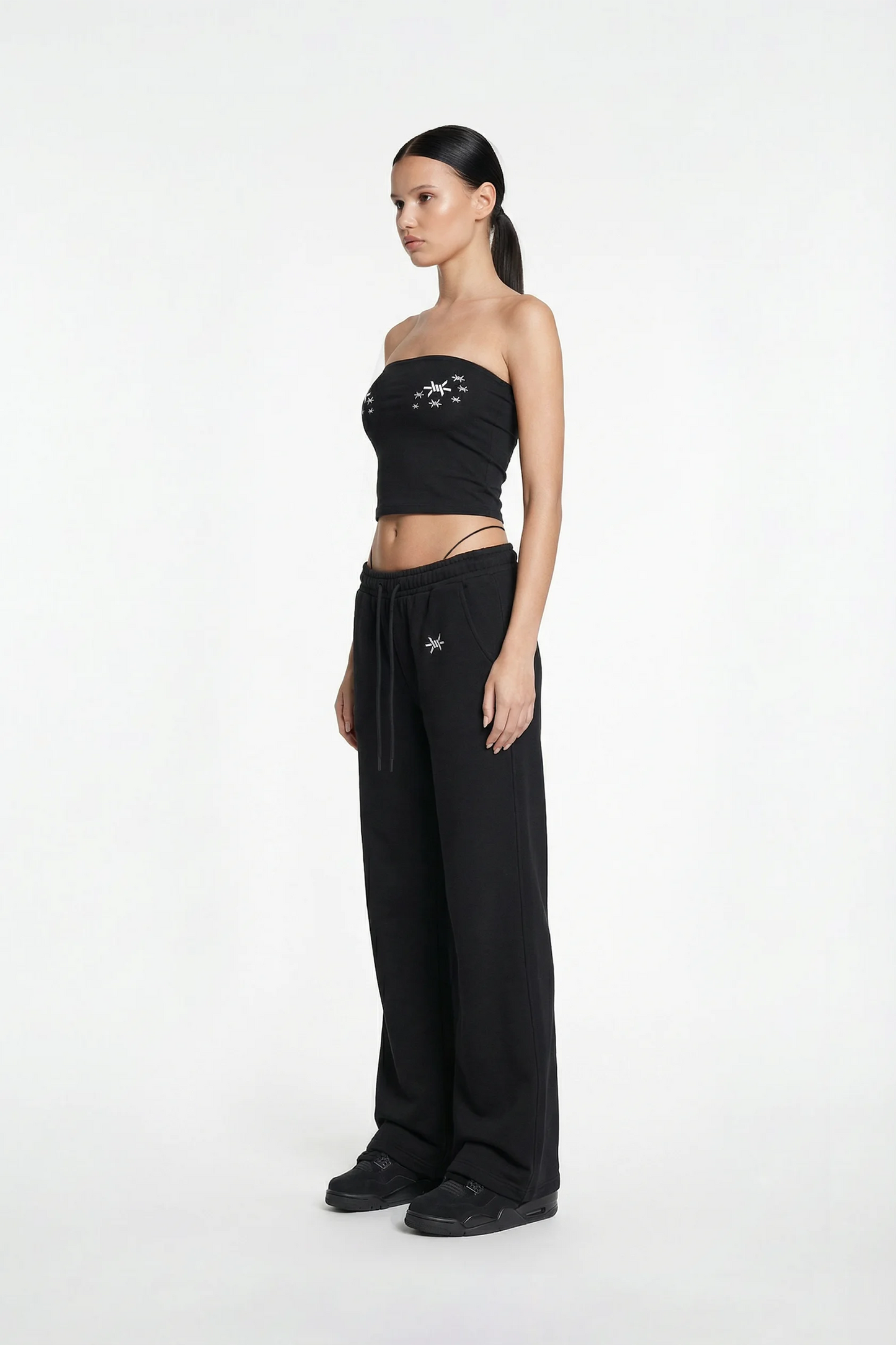 Woman wearing a black strapless top and wide-leg pants with brand logos on a white background