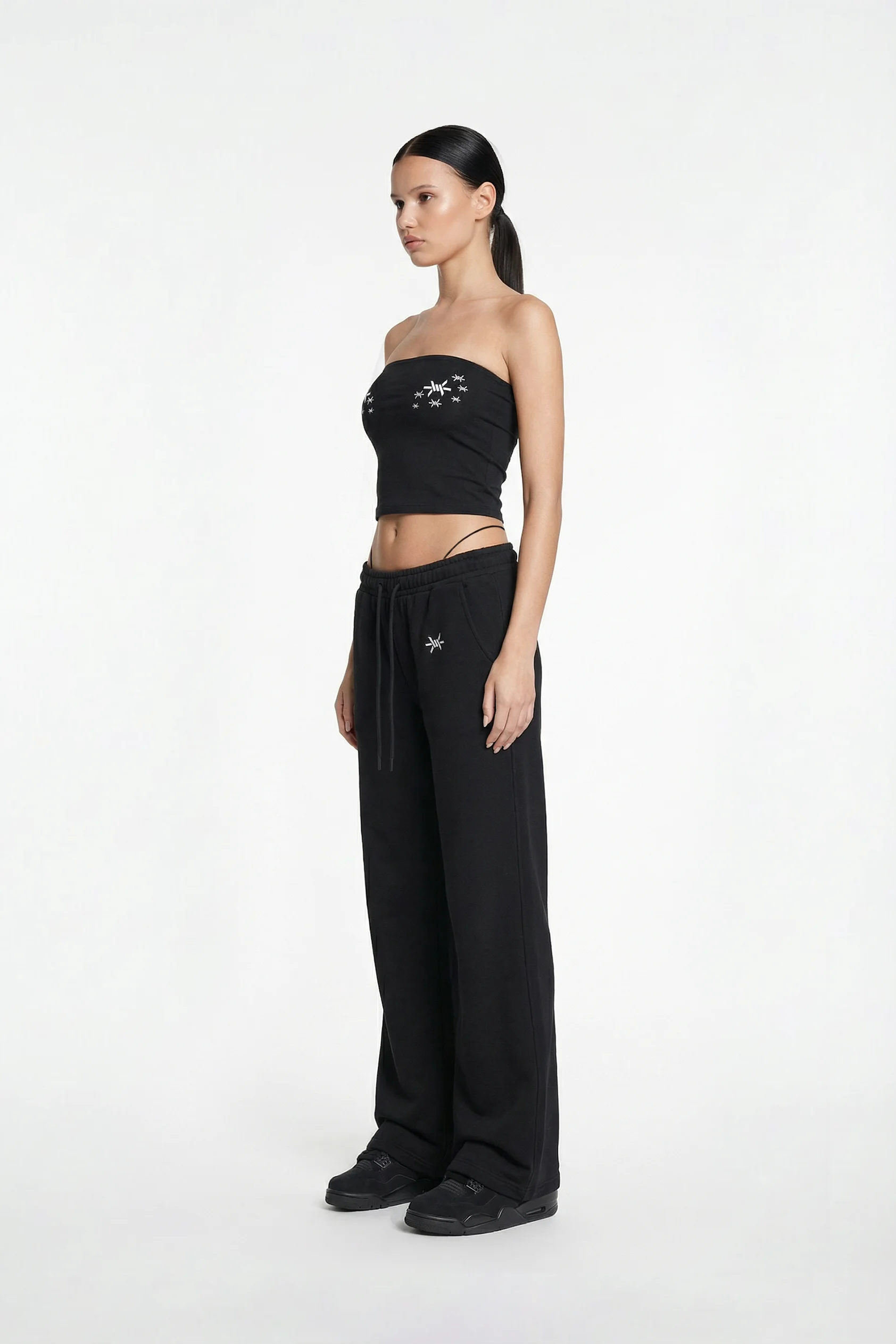 Woman wearing a black strapless top and wide-leg pants with brand logos on a white background